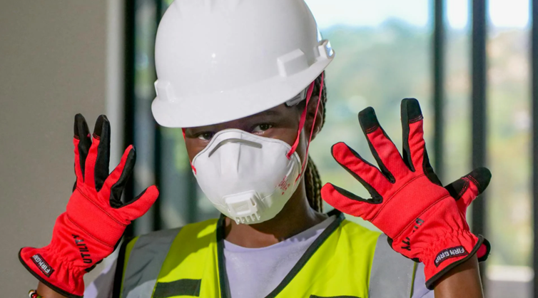Vrouw met witte helm, stofmasker en rode werkhandschoenen toont haar PBM op een bouwlocatie