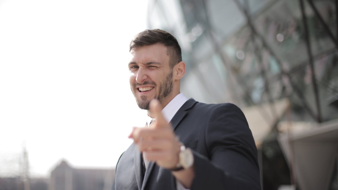 Man in pak knipoogt en wijst zelfverzekerd naar de camera – hét voorbeeld van “je van het”-uitstraling.