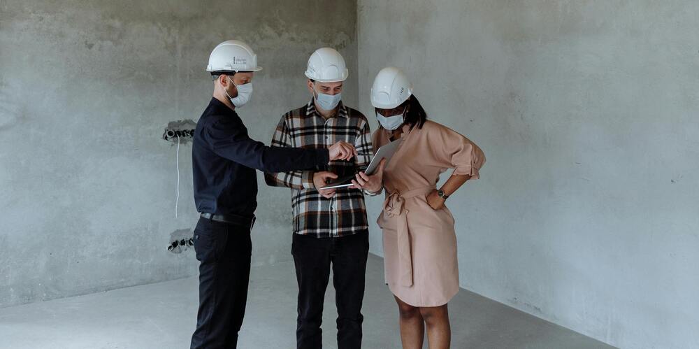 Drie bouwprofessionals met helmen en mondkapjes bekijken samen een tablet in een lege ruimte